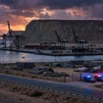 Editorial illustration of Gwadar port at dusk with cargo cranes silhouetted against a dramatic sky, a coastal highway in the foreground, a fenced construction camp, and security vehicles with flashing blue and red lights parked nearby, with rugged mountains behind.