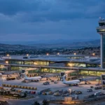Modern airport terminal at dusk with control tower, aircraft parked at gates and cargo containers on the apron