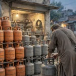 Stacks of orange and grey LPG gas cylinders outside a small neighbourhood shop at dusk, with a shopkeeper seen from behind checking a cylinder