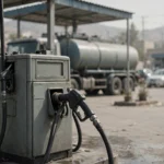 Fuel pump with nozzle in the foreground and a tanker truck at a fuel station in daylight