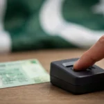 A finger pressing a biometric fingerprint scanner beside a blurred green Pakistani identity card on a desk, with the Pakistani flag out of focus in the background
