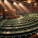 Empty Senate chamber with rows of green desks and microphones, Pakistani flags lined along a wood-panelled wall, and sunlight streaming through tall windows.
