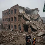 Rescue workers with a stretcher outside the collapsed brick school building in Peshawar at dawn, with emergency vehicles and the Pakistani flag nearby, after the Al-Khair school roof collapse in October 2026