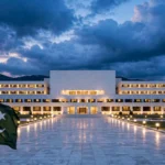 An editorial illustration of Pakistan's Parliament House in Islamabad at dusk, with the Pakistani national flag waving in the foreground under a dramatic cloudy sky, October 2026