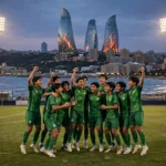 A youth football team in green jerseys celebrating on a floodlit stadium pitch at dusk with Baku's Flame Towers skyline and the Caspian Sea in the background, October 2026.