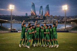 A youth football team in green jerseys celebrating on a floodlit stadium pitch at dusk with Baku's Flame Towers skyline and the Caspian Sea in the background, October 2026.