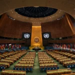 A wide interior view of the United Nations General Assembly hall with rows of delegate desks, the UN emblem on screens at the front and the Pakistani national flag among the national flags, October 2026