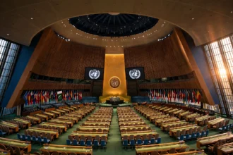 A wide interior view of the United Nations General Assembly hall with rows of delegate desks, the UN emblem on screens at the front and the Pakistani national flag among the national flags, October 2026