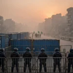 Police in riot gear behind barricades and shipping containers on a road at dawn as hundreds of PTI members are detained in Khyber Pakhtunkhwa after the emergency in October 2026
